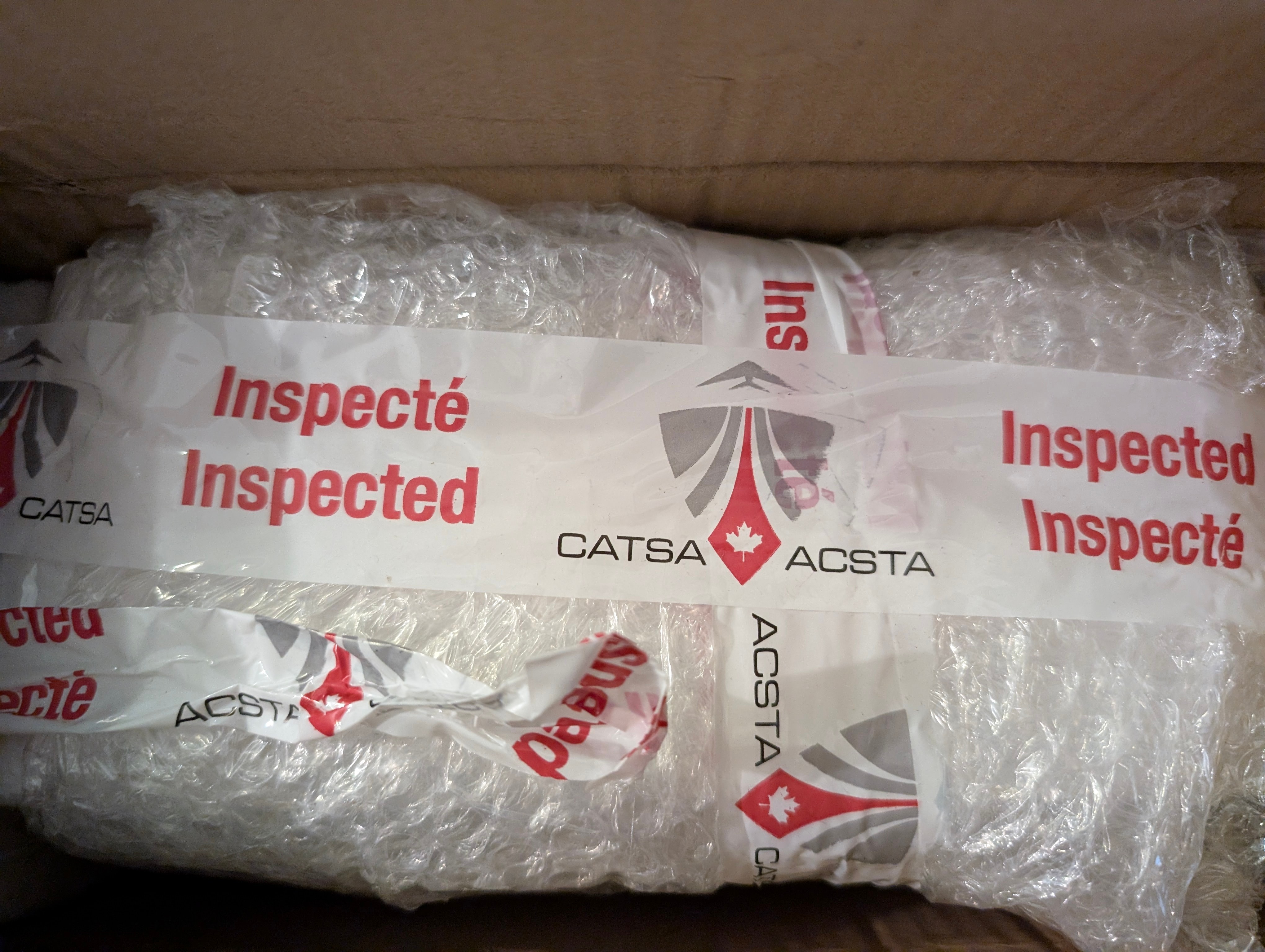A close-up of an item inside a cardboard box, wrapped in bubble wrap and sealed with security tape. The tape reads "Inspected / Inspecté" in red text and features the CATSA (Canadian Air Transport Security Authority) and ACSTA logos, which include a maple leaf and a stylized shield. The tape is slightly crumpled, indicating the package was opened and re-sealed after inspection.